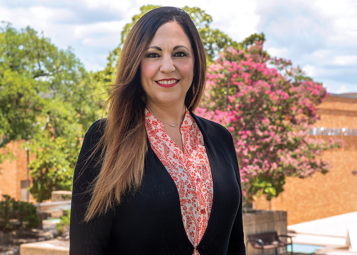 Anne Gaillard stands in the shade on the SHSU campus with the flowers and a fountain in the background.